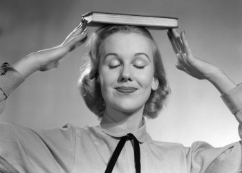 1950s BLOND WOMAN SMILING WITH EYES CLOSED BALANCING BOOK ON HER HEAD  (Photo by Debrocke/ClassicStock/Getty Images)