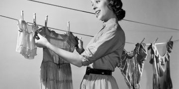 1950s SMILING WOMAN HOUSEWIFE HANGING WASH CHILD DRESS ON CLOTHESLINE TO DRY  (Photo by Debrocke/ClassicStock/Getty Images)